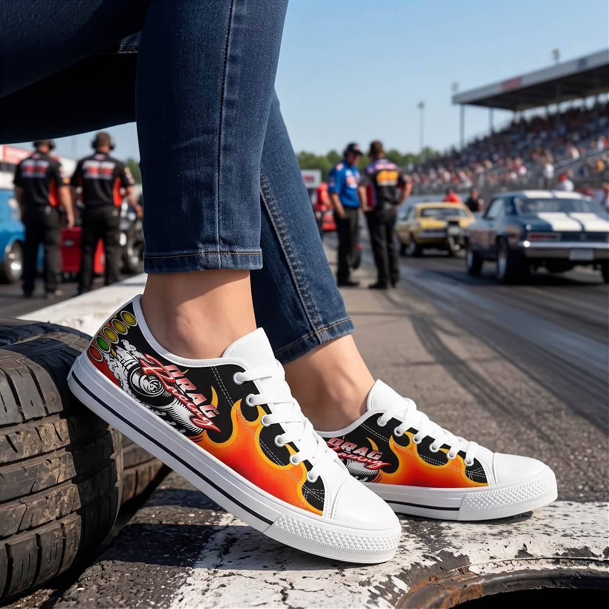 White low-top canvas sneakers with a vibrant orange and yellow flame graphic and a hot rod illustration on the side, worn by a person sitting on a tire at a racetrack.