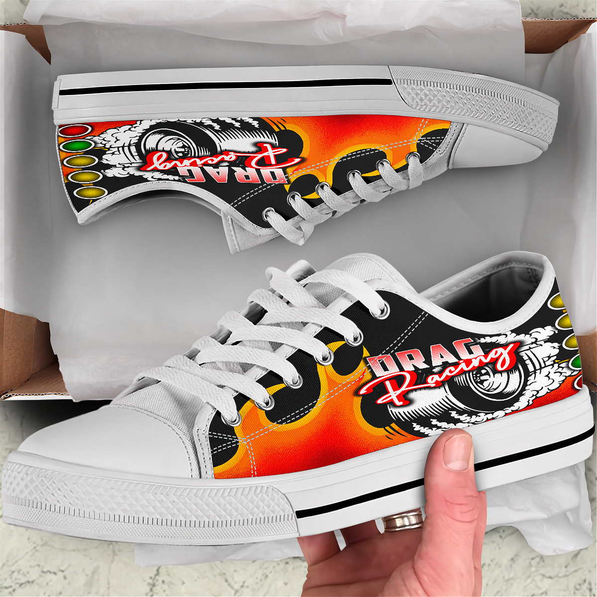A pair of white low-top sneakers with a vibrant orange, yellow, and red flame graphic and 'DRAG Racing' text on the sides, shown with one shoe in its box and the other held by a hand.