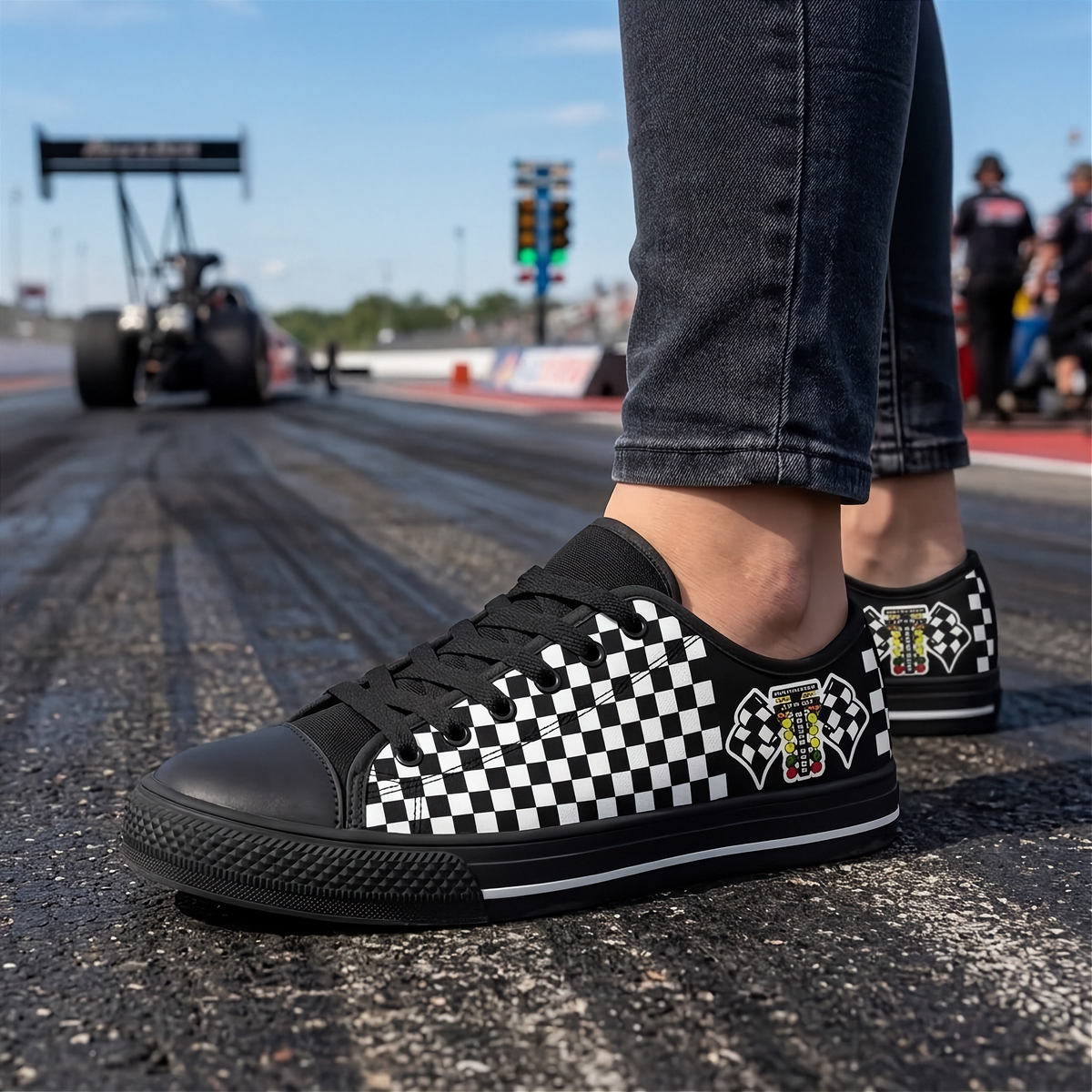 A person wearing black and white checkered low-top sneakers on an asphalt racetrack, with the rear of a race car and track barriers blurred in the background.