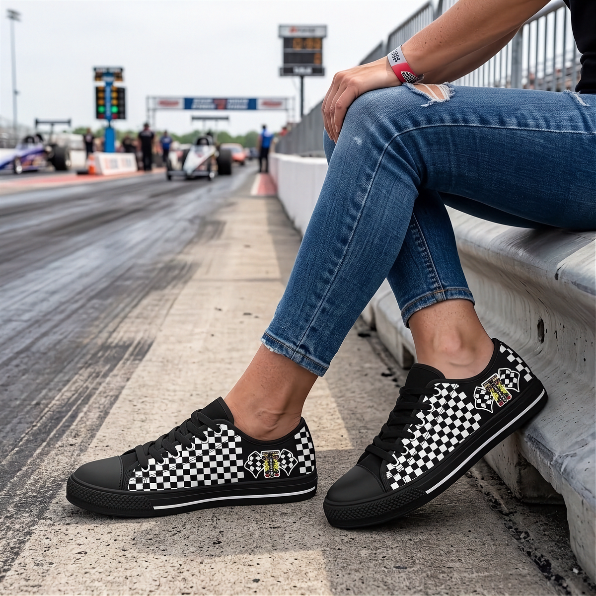 A person sitting on a concrete barrier at a racetrack wearing black low-top sneakers with a black and white checkered pattern and a small yellow-and-black crest patch on the side.