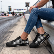 A person sitting on a concrete barrier at a racetrack wearing black low-top sneakers with a black and white checkered pattern and a small yellow-and-black crest patch on the side.