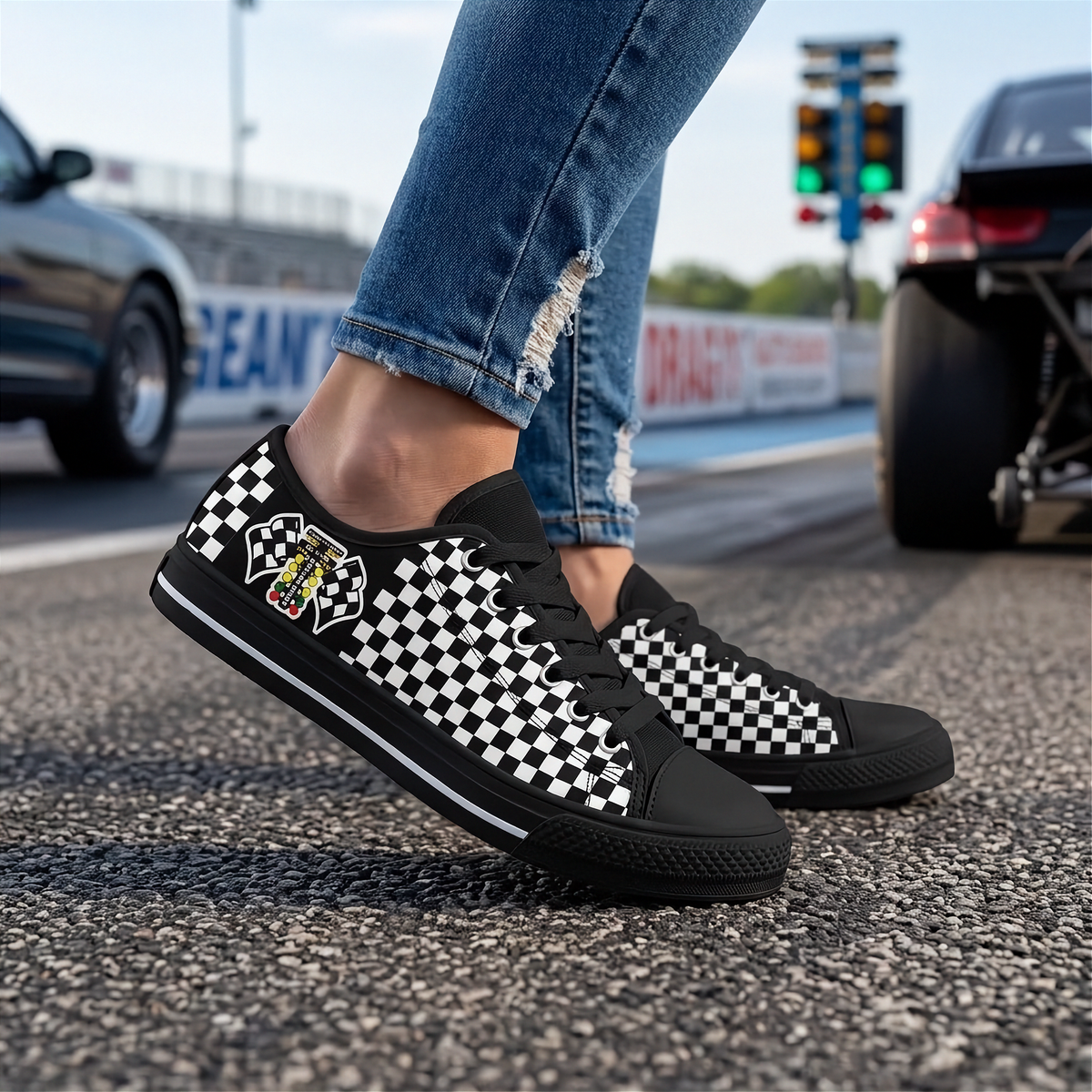 A person wearing black low-top sneakers with a black-and-white checkerboard pattern and a small racing crest on the side, standing on an asphalt racetrack with cars and traffic lights in the background.