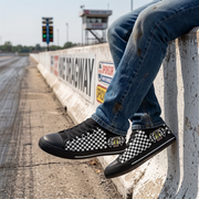 A person sitting on a concrete barrier at a racetrack wearing black low-top sneakers with a black-and-white checkerboard pattern and small racing decals, along with blue jeans.