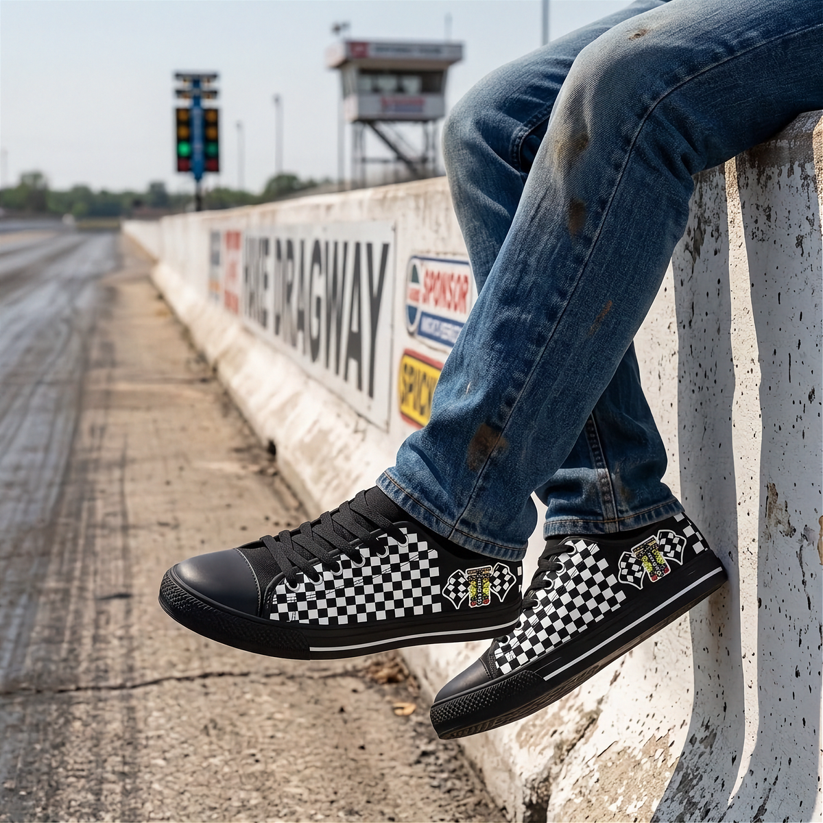 A person sitting on a concrete barrier at a racetrack wearing black low-top sneakers with a black-and-white checkerboard pattern and small racing decals, along with blue jeans.