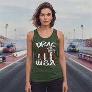 A woman stands on a drag racing track wearing an olive green racerback tank top with a graphic that reads 'DRAG Racing USA' in white over a red, white, and green American flag design. Racing cars and the racetrack are visible in the background.