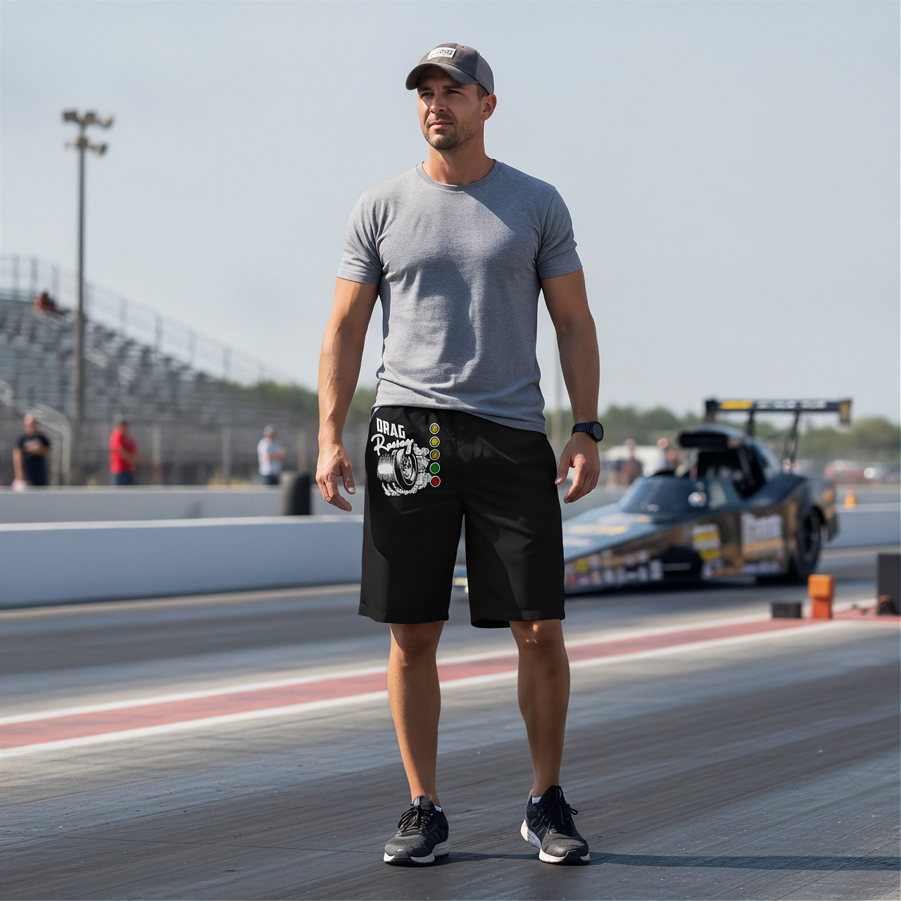 A man standing on a drag racing track wearing a gray t-shirt, black shorts with a white and green graphic print, and black sneakers. A drag race car is visible in the background.