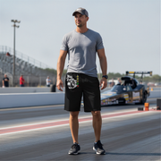 A man standing on a drag racing track wearing a gray t-shirt, black shorts with a white and green graphic print, and black sneakers. A drag race car is visible in the background.