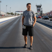 A man stands on a drag racing track wearing a gray crewneck t-shirt and black athletic shorts with a white graphic print on the left leg. The background shows the racetrack, starting line, and a control tower under a clear sky.