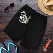 Black athletic shorts with a 'DRAG Racing' tire graphic and traffic light design, styled in a flat lay on a dark wood background with sunglasses, a straw hat, flip-flops, and a watch.