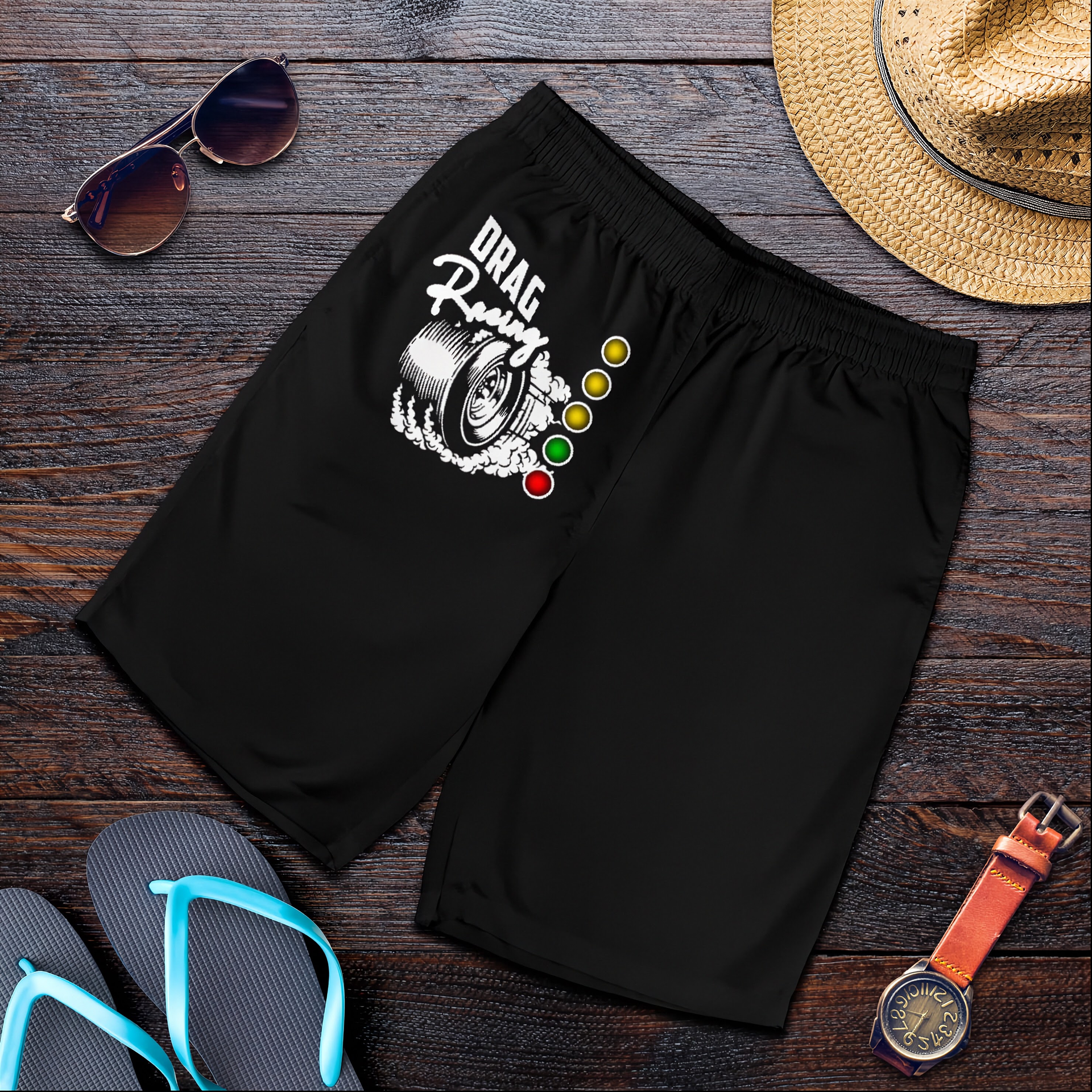 Black athletic shorts with a 'DRAG Racing' tire graphic and traffic light design, styled in a flat lay on a dark wood background with sunglasses, a straw hat, flip-flops, and a watch.