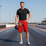 A man stands on a racetrack wearing a black t-shirt and bright red athletic shorts with a drag racing graphic print on the left leg, holding a black baseball cap in one hand.