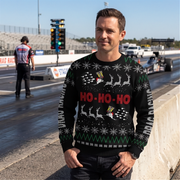 A man standing on a drag race track wearing a black Christmas sweater with red 'HO-HO-HO' text, white reindeer, and green snowflake patterns, with race cars and a track environment in the background.