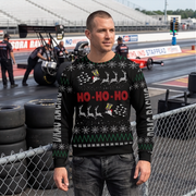 A man wearing a black Christmas sweater with a festive holiday pattern stands in front of a chain-link fence at a drag racing track. The sweater features 'HO-HO-HO' in red text, white reindeer silhouettes, snowflakes, and green accents.