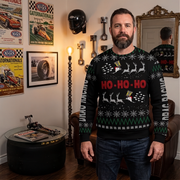 A man wearing a black Christmas sweater with a festive holiday pattern and 'HO-HO-HO' text, standing in a themed room with racing memorabilia.