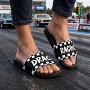 A person wearing black slide sandals with a black-and-white checkered border and the words 'DRAC RACING' printed across the straps, standing on a drag strip with cars and track signage in the background.