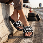 A person sitting on a concrete barrier at a racetrack wearing black slide sandals with a white checkerboard pattern and the words 'DRAG RACING' across the straps.