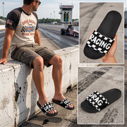 A man wearing black and white checkered slide sandals with 'RACING' printed on the straps, sitting on a concrete racetrack barrier. The image includes two inset close-ups showing the sandals from different angles, one held in a hand and one on the racetrack pavement.
