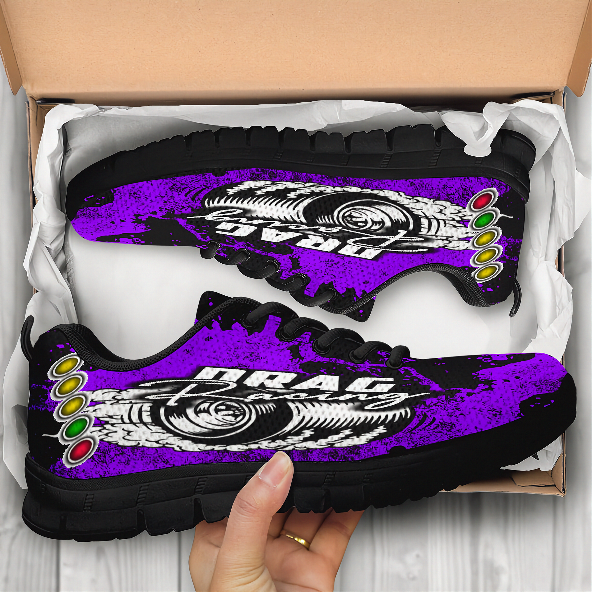 Purple and black sneakers with 'Drag' text held by a hand, displayed in an open cardboard box.