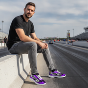 Man sitting on a barrier at a race track wearing black and purple sneakers.