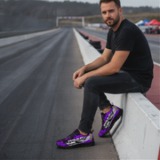 Man sitting on a race track wall wearing purple sneakers with black patterns.