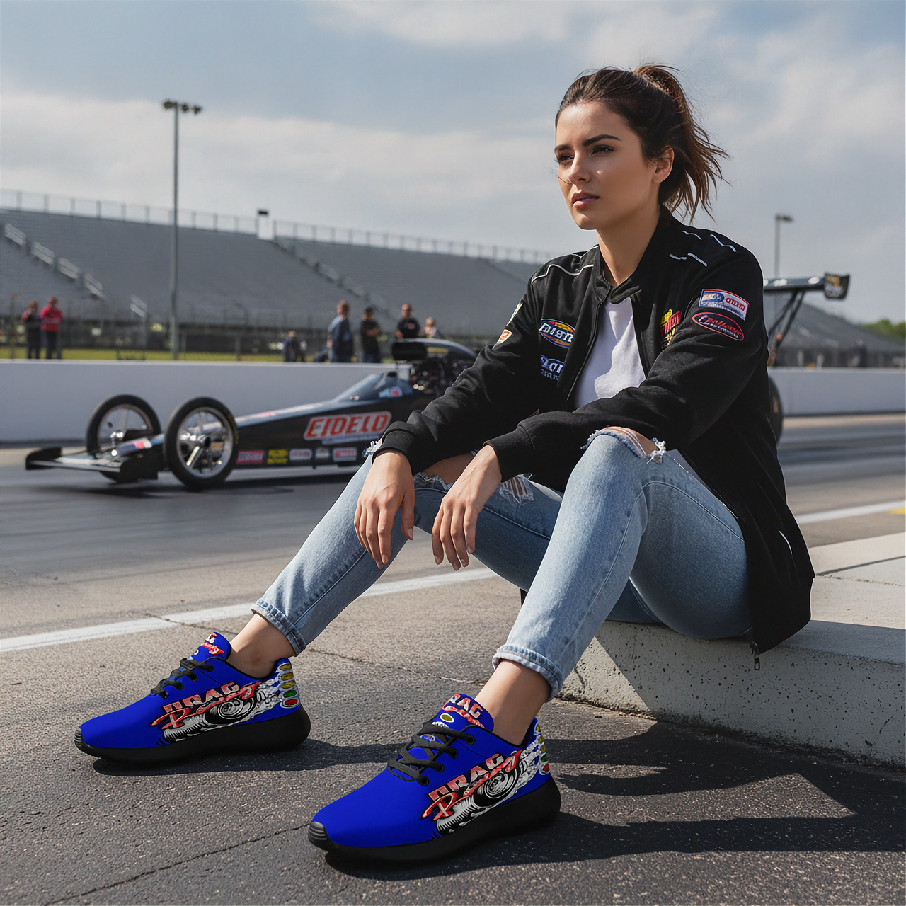 A woman sits on a racetrack curb wearing bright blue and red drag racing sneakers with 'Ryde' branding, a black bomber jacket, and light blue jeans, with a drag race car and grandstands in the background.