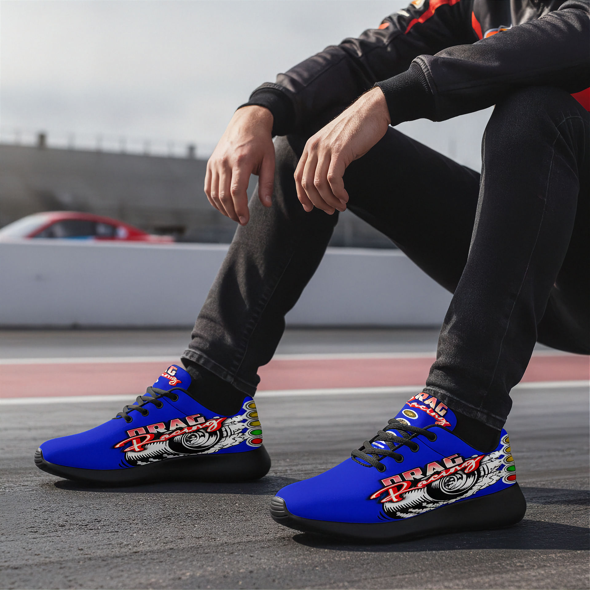 A person sitting outdoors wearing bright blue low-top sneakers with a large 'DRAG' graphic and racing-style wheel illustration on the sides, paired with black jeans and a black sweatshirt.