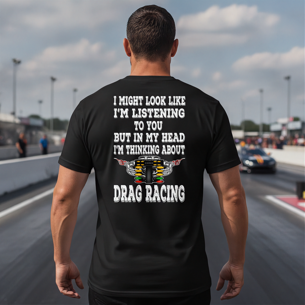 A man viewed from behind wearing a black t-shirt with a white and multicolor graphic print about drag racing, standing on a racetrack.