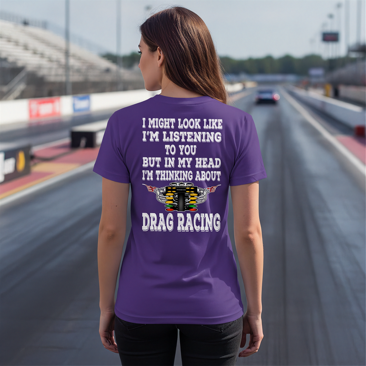 A woman stands on a drag racing track wearing a purple t-shirt with a humorous drag racing graphic and text on the back, viewed from behind.