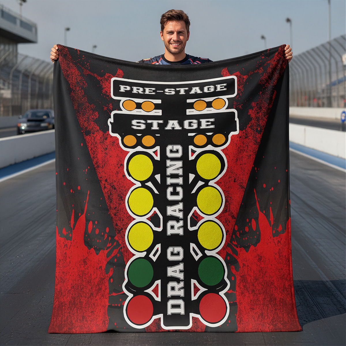 A man stands on a racetrack holding up a large printed blanket or towel featuring a drag racing theme with red, black, yellow, green, and white graphics.