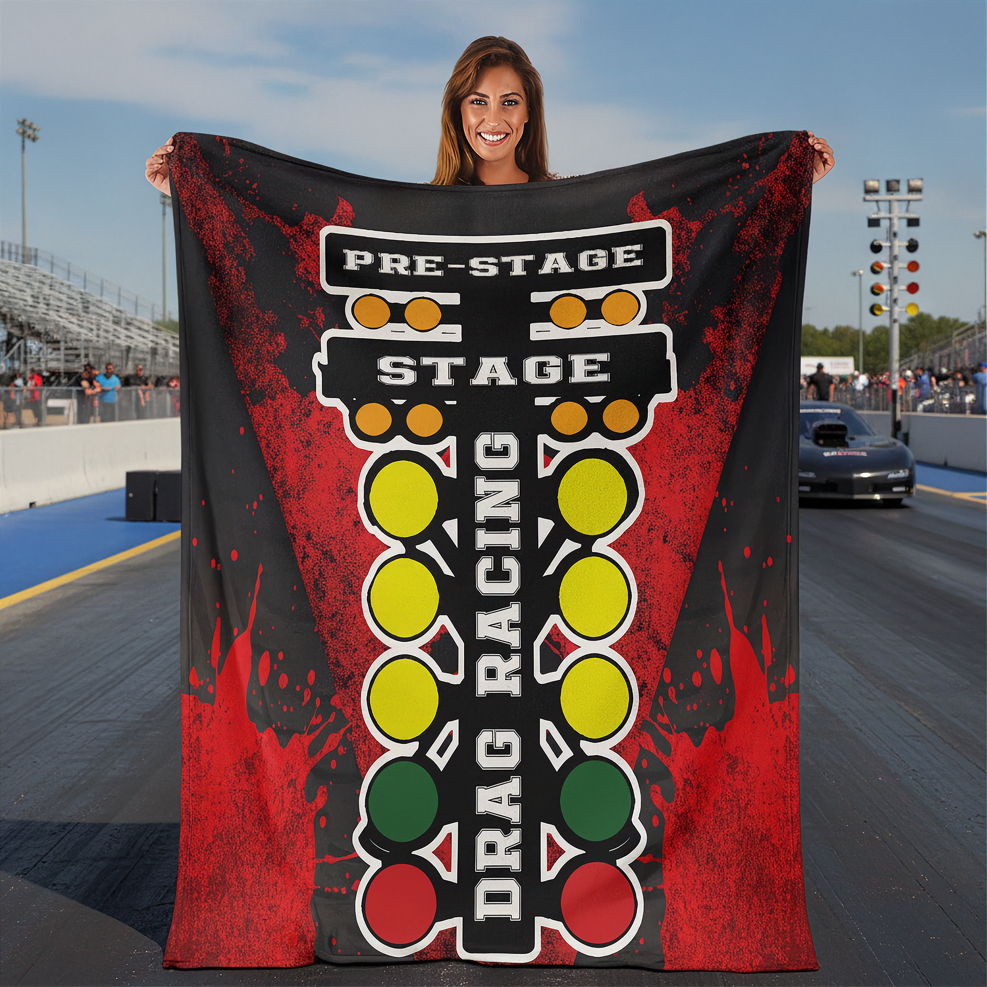 A woman stands on a drag racing track holding up a large black and red graphic blanket or towel featuring a vertical drag racing lights design with yellow, green, and red circles and the words 'PRE-STAGE', 'STAGE', and 'DRAG RACING'.