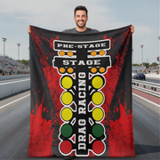 A man standing on a racetrack holds up a large black and red drag racing themed blanket or towel featuring a vertical light bar design with yellow, green, and red lights and the words 'PRE-STAGE', 'STAGE', and 'DRAG RACING'.