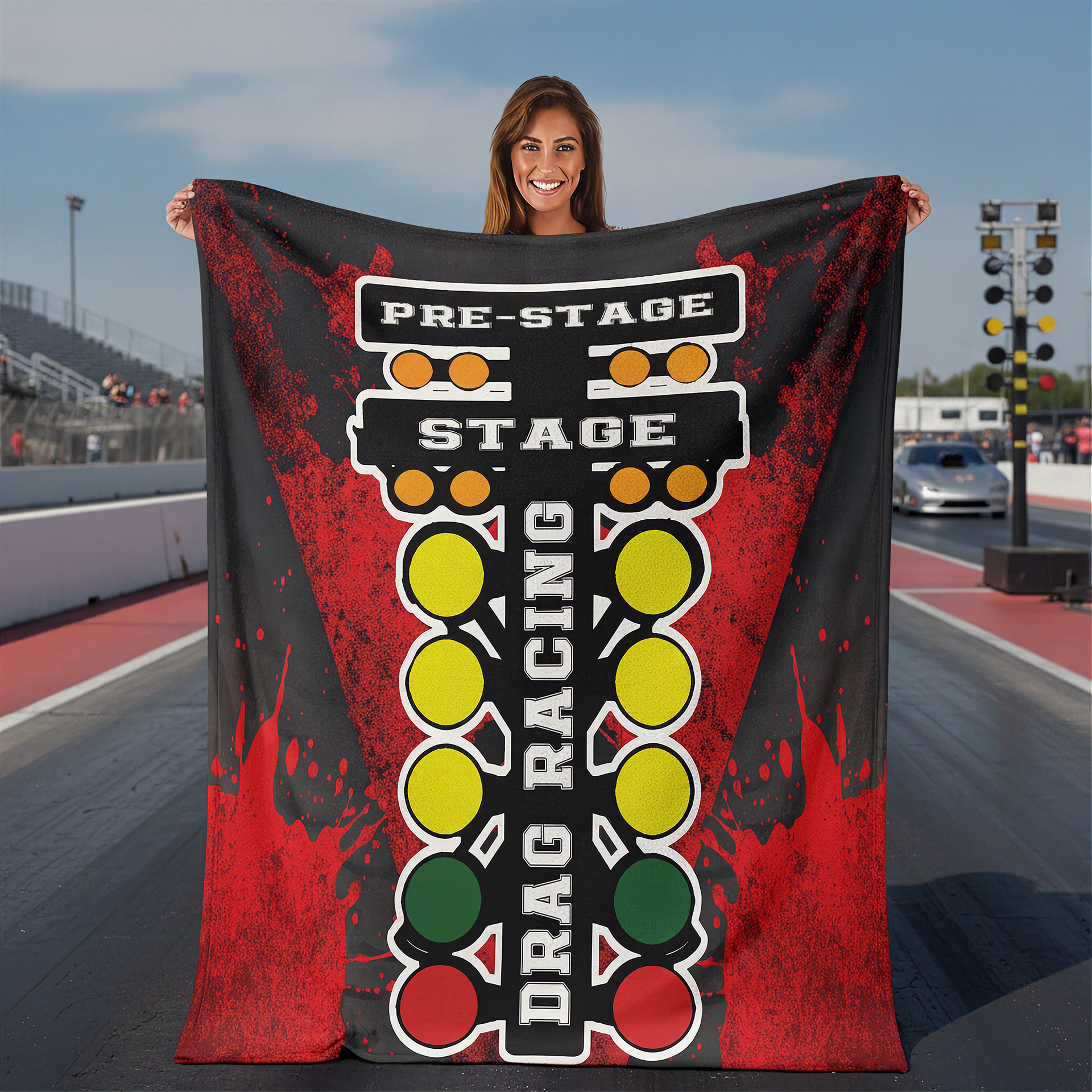 A woman stands on a racetrack holding up a large printed blanket or tapestry featuring a drag racing theme with the words 'PRE-STAGE', 'STAGE', and 'DRAG RACING' over a traffic-light-style graphic in red, yellow, green, and white.