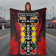 A woman stands on a racetrack holding up a large printed blanket or tapestry featuring a drag racing theme with the words 'PRE-STAGE', 'STAGE', and 'DRAG RACING' over a red, black, yellow, green, and white graphic.