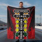 A man standing on a racetrack holds up a large printed blanket or tapestry featuring a drag racing theme with a red and black background, white text reading 'PRE-STAGE', 'STAGE', and 'DRAG RACING', and yellow, green, and red circular lights.