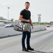 A man stands on a racetrack holding a gray duffel bag with black straps and white racing graphics. The bag features a circular tire graphic and a row of red, yellow, and green oval accents on the front.
