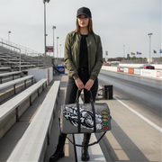 A woman stands on a racetrack holding a gray duffel bag with a large black and white skull graphic and the words 'Oral Roberts'. The racetrack, bleachers, and distant cars are visible in the background.