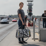 A woman stands on a racetrack holding a gray duffel bag with a black and white racing graphic and colored oval accents. The background shows a blurred race track, cars, and track crew.