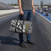 A man standing on a racetrack holding a gray duffel bag with a black and white graphic design and red, yellow, and green oval accents. The bag has 'Drain Race' printed on it.