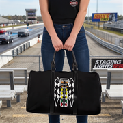 A person standing at a racetrack holding a black duffel bag with a large racing-themed patch on the front. The patch features checkered flags, a vertical racing flag graphic, and the word 'DRAG'.RACING