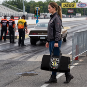 A woman stands on a racetrack holding a black duffel bag with a large DRAG RACING graphic. A race car, track officials, and track signage are visible in the background.