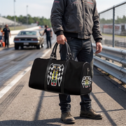 A person standing on a racetrack holds a black duffel bag with racing graphics, including a checkered flag motif and a stylized figure.