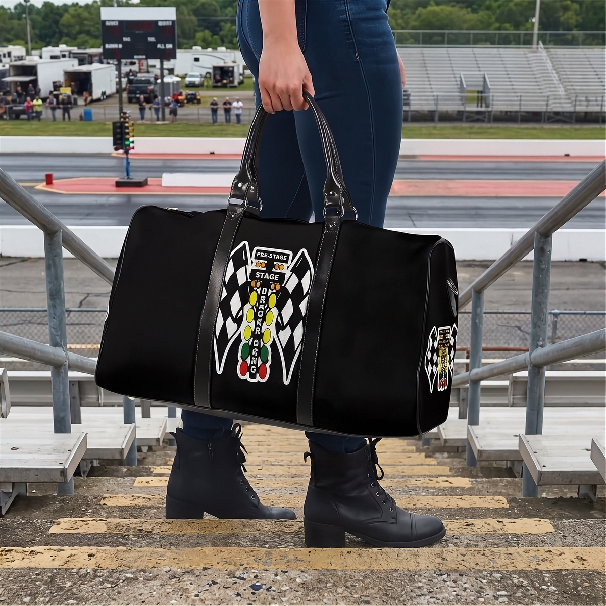 A person standing on race track bleachers holds a black duffel bag with a large drag  racing graphic featuring checkered flags and a yellow-and-black driver illustration. The background shows the racetrack, grandstands, and infield.