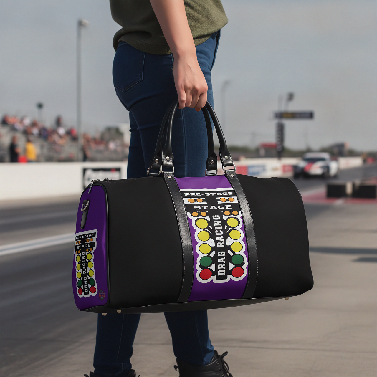 A person standing on a racetrack holds a black duffel bag with purple side panels and a large racing-style graphic reading 'PRE-STAGE', 'STAGE', and 'DRAG RACING'.