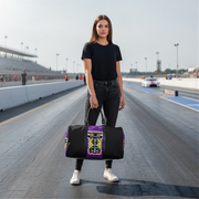 A woman stands on a racetrack holding a black duffel bag with a purple side panel and a colorful drag racing graphic print. The racetrack, grandstands, and distant race cars are visible in the background.