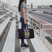A woman stands on outdoor stadium bleachers holding a black duffel bag with a bold purple and multicolor drag racing graphic panel on the front.