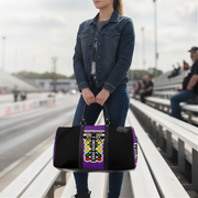 A woman standing on stadium bleachers holds a black duffel bag with a purple side panel and a colorful racing-style graphic. The background shows a blurred racetrack and seated spectators.
