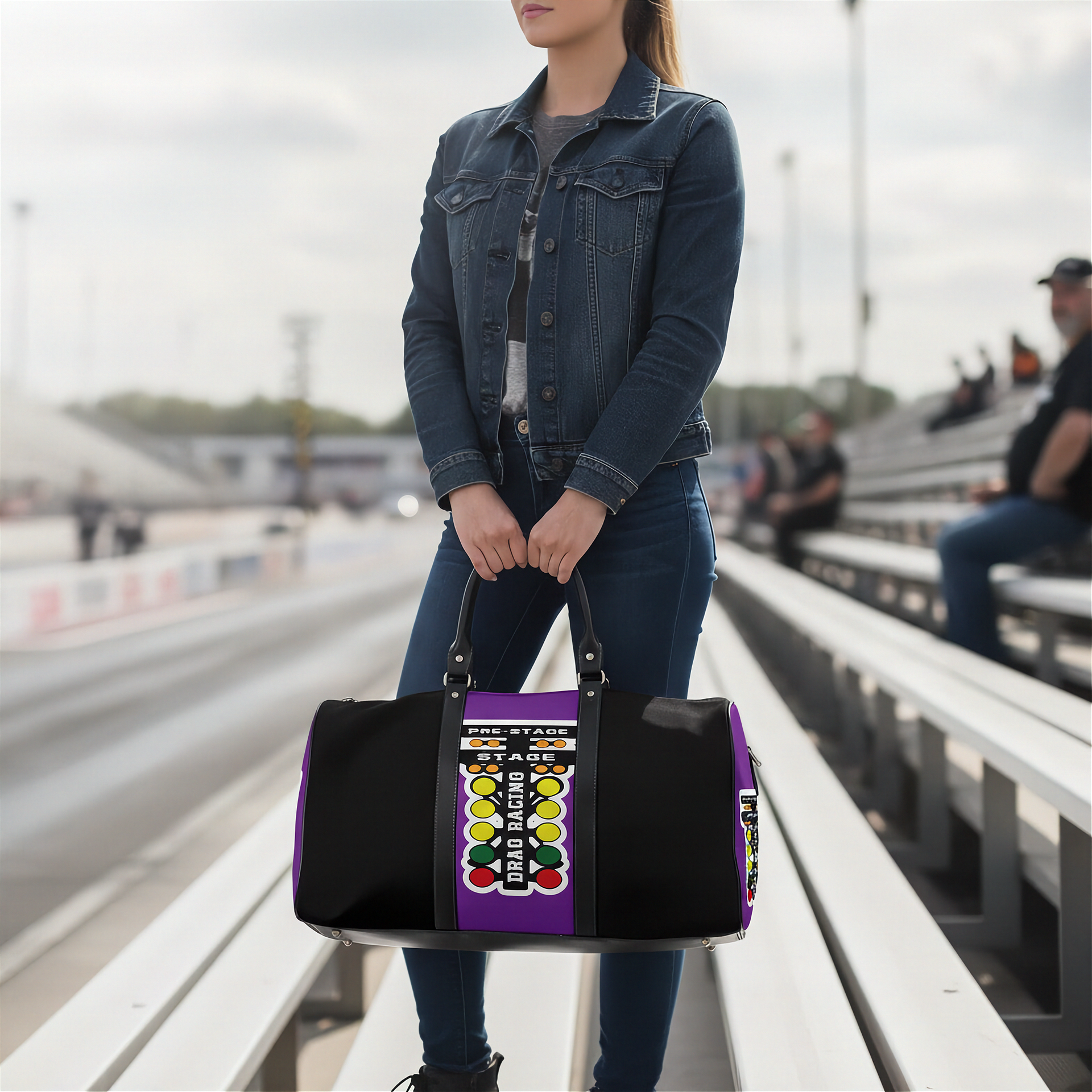 A woman standing on stadium bleachers holds a black duffel bag with a purple side panel and a colorful racing-style graphic. The background shows a blurred racetrack and seated spectators.