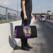 A person standing on a racetrack holds a black duffel bag with purple side panels and a large colorful drag racing tree graphic on the front.