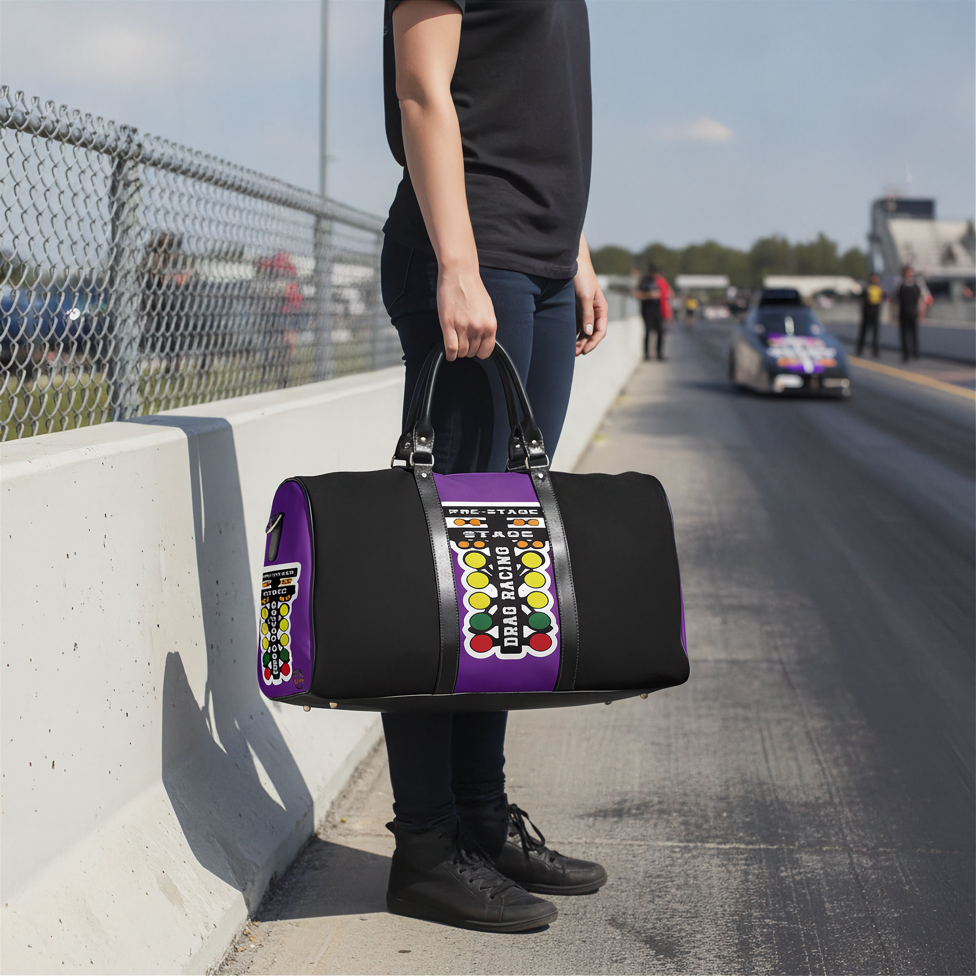 A person standing on a racetrack holds a black duffel bag with purple side panels and a large colorful drag racing tree graphic on the front.