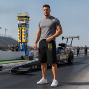A muscular man stands on a drag racing track wearing a gray t-shirt, black shorts with a colorful graphic on the side, and white sneakers, with a drag race car and a tower in the background.